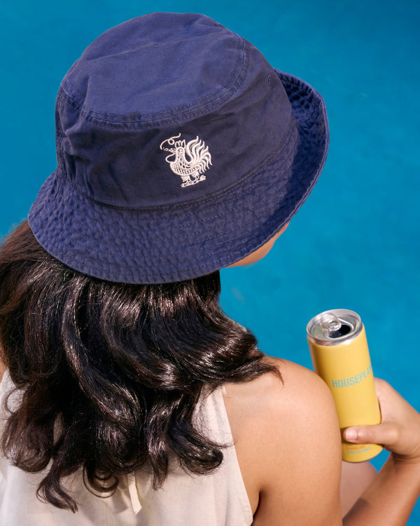 Person wearing a navy bucket hat with a logo, holding a yellow can against a blue background