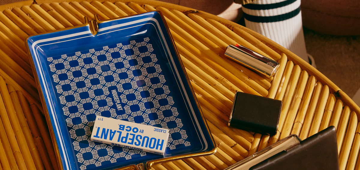 A blue and white patterned Houseplant ashtray with gold trim, styled on a yellow woven rattan table alongside a silver lighter, a black case, and a notebook.