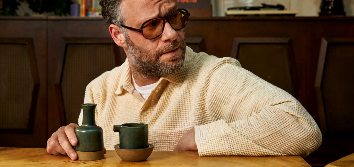 Seth Rogen seated at a wooden table beside a dark green ashtray set